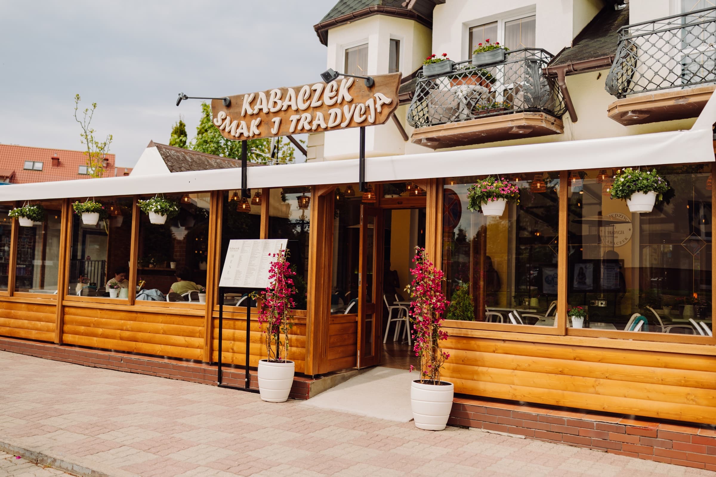 Restaurant Kabaczek in Ustronie Morskie von der Straße aus — Holzschild, blühende Terrasse und blauer Himmel